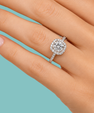A close-up of a hand wearing a halo ring with a round brilliant-cut center stone and a pavé-set band, shown against a solid light blue background.