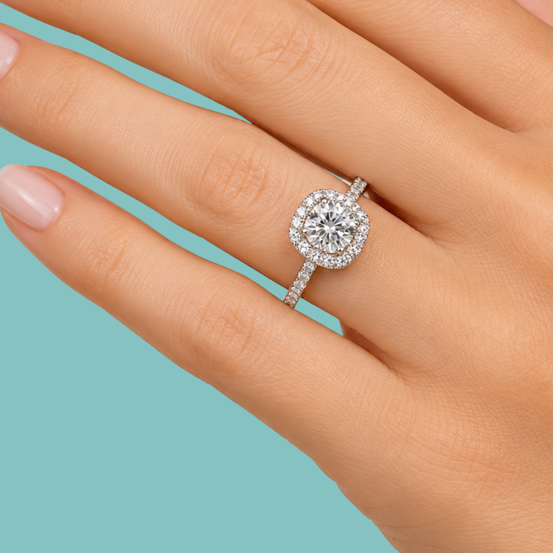 A close-up of a hand wearing a halo ring with a round brilliant-cut center stone and a pavé-set band, shown against a solid light blue background.