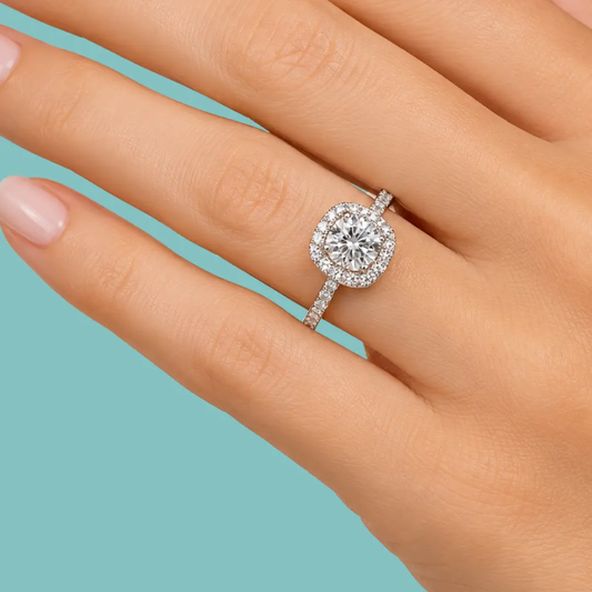 A close-up of a hand wearing a halo ring with a round brilliant-cut center stone and a pavé-set band, shown against a solid light blue background.