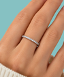 A close-up of a hand wearing a thin silver eternity ring set with small clear stones, against a solid light blue background.