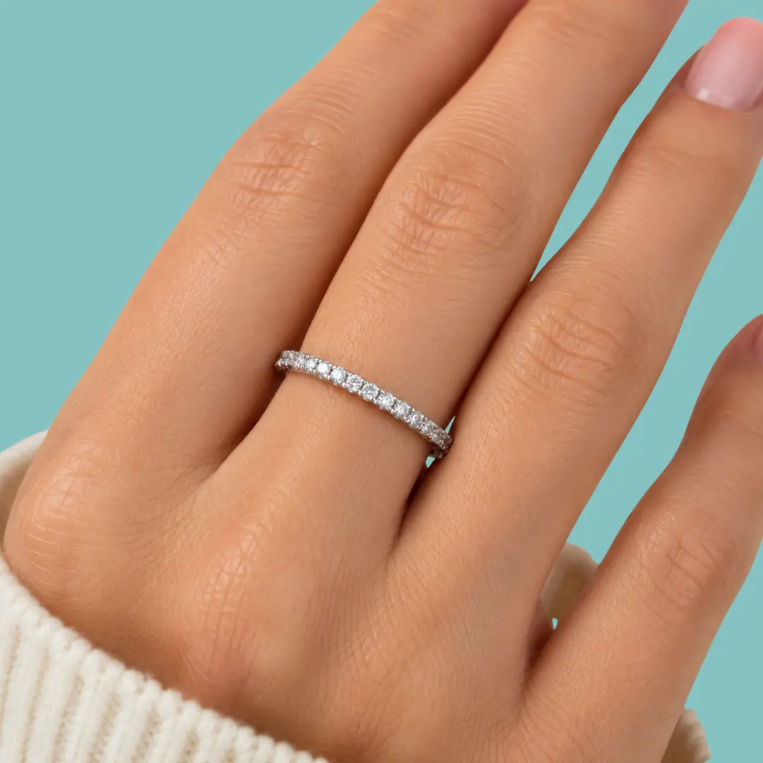 A close-up of a hand wearing a thin silver eternity ring set with small clear stones, against a solid light blue background.