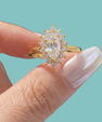 A gold ring with a marquise-cut clear center stone and surrounding round stones, held between fingers against a solid turquoise background.