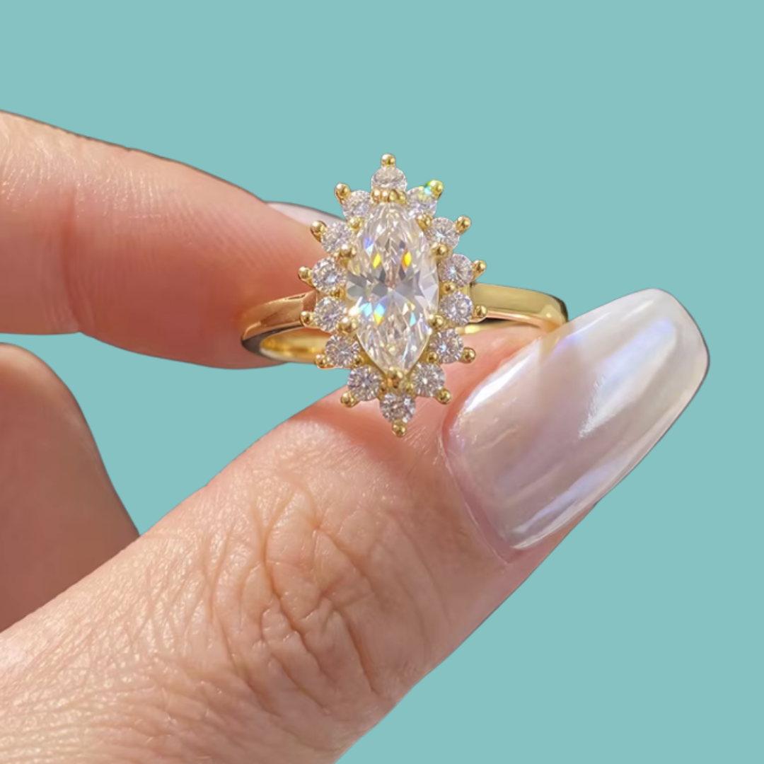 A gold ring with a marquise-cut clear center stone and surrounding round stones, held between fingers against a solid turquoise background.