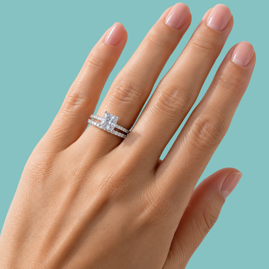 A close-up of a hand wearing a silver-toned double ring set with a large square-cut clear center stone and a matching pavé band, against a solid light aqua background.