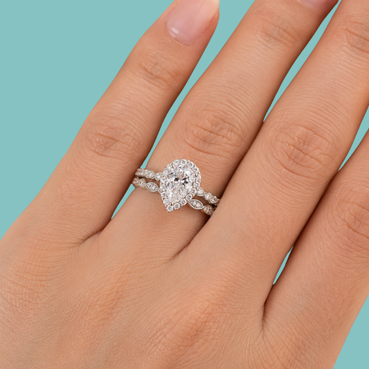 A close-up of a hand wearing a silver-toned ring set with a pear-cut diamond and smaller diamonds along the band, against a solid light blue background.