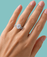 A close-up of a hand wearing a double-band engagement ring with a round brilliant-cut center stone and pavé bands, set against a solid light teal background.