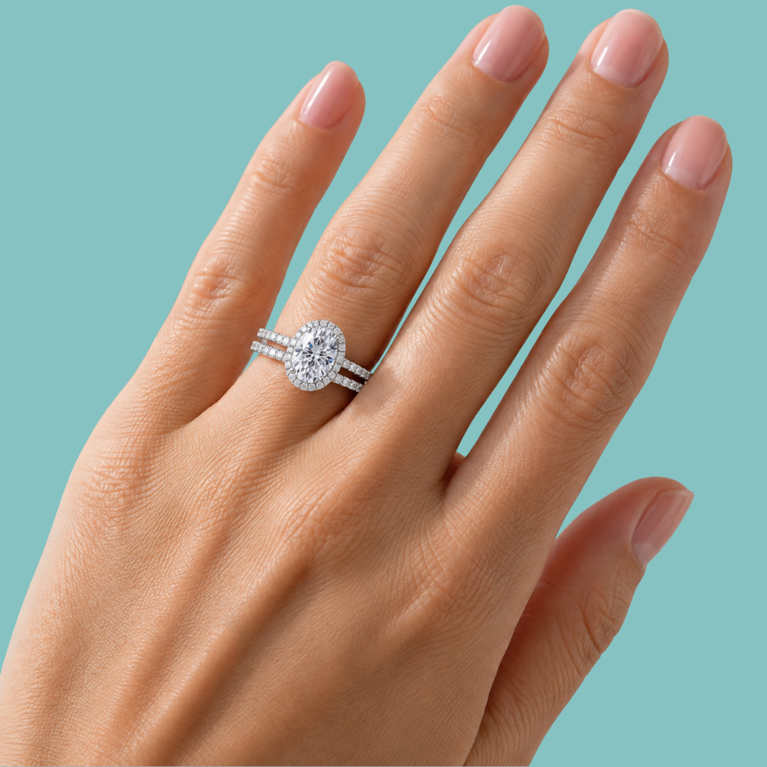 A close-up of a hand wearing a double-band engagement ring with a round brilliant-cut center stone and pavé bands, set against a solid light teal background.