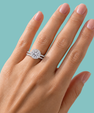 A close-up of a hand wearing a diamond halo engagement ring with a matching pave band, set against a solid teal background.