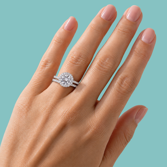A close-up of a hand wearing a diamond halo engagement ring with a matching pave band, set against a solid teal background.