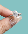 A close-up of a hand with manicured nails holding a silver-toned solitaire engagement ring with an oval-cut clear gemstone against a solid light teal background.