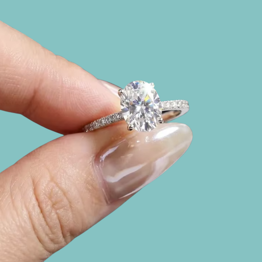 A close-up of a hand with manicured nails holding a silver-toned solitaire engagement ring with an oval-cut clear gemstone against a solid light teal background.