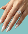 A close-up of a hand with long, glossy nude manicured nails wearing a sparkling diamond engagement ring on a solid teal background.
