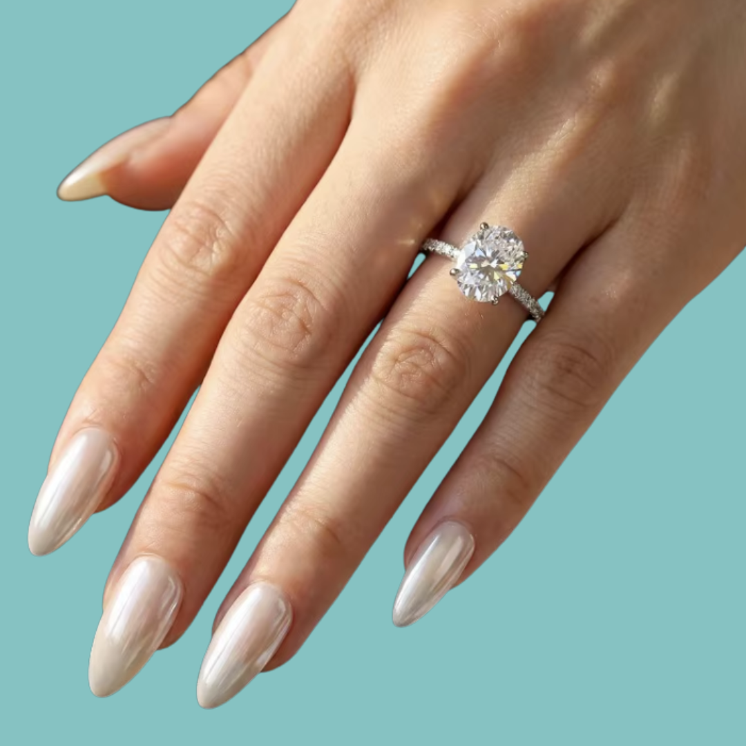 A close-up of a hand with long, glossy nude manicured nails wearing a sparkling diamond engagement ring on a solid teal background.