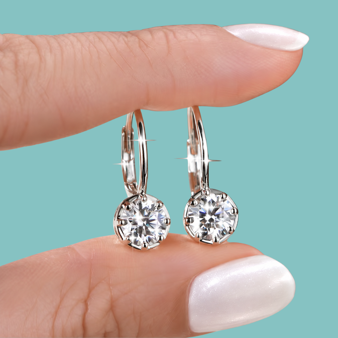 A close-up of a person's hand with white manicured nails holding a pair of silver hoop earrings with round-cut clear gemstones against a solid teal background.