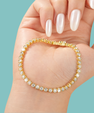 A gold tennis bracelet with clear round crystals, shown worn on a hand with long white manicured nails against a solid light blue background.