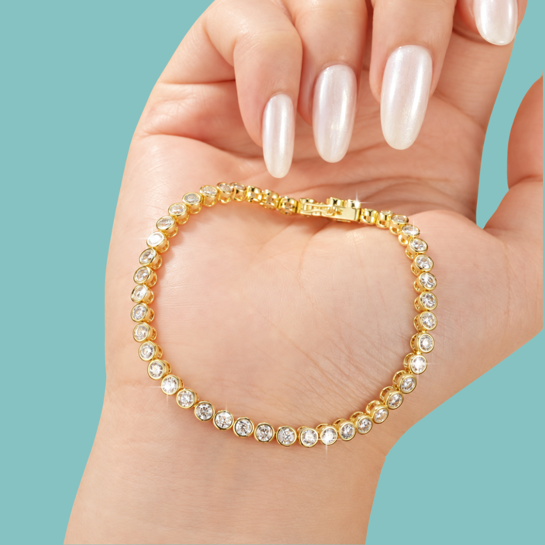 A gold tennis bracelet with clear round crystals, shown worn on a hand with long white manicured nails against a solid light blue background.