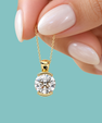 A close-up of a hand with pale pink manicured nails holding a delicate gold chain with a round-cut clear gemstone pendant in a gold bezel setting, set against a solid light teal background.