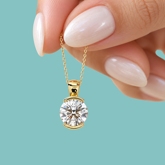 A close-up of a hand with pale pink manicured nails holding a delicate gold chain with a round-cut clear gemstone pendant in a gold bezel setting, set against a solid light teal background.
