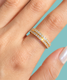 A close-up of a hand wearing a double-row yellow gold eternity ring set with clear pavé gemstones, set against a solid light blue background.