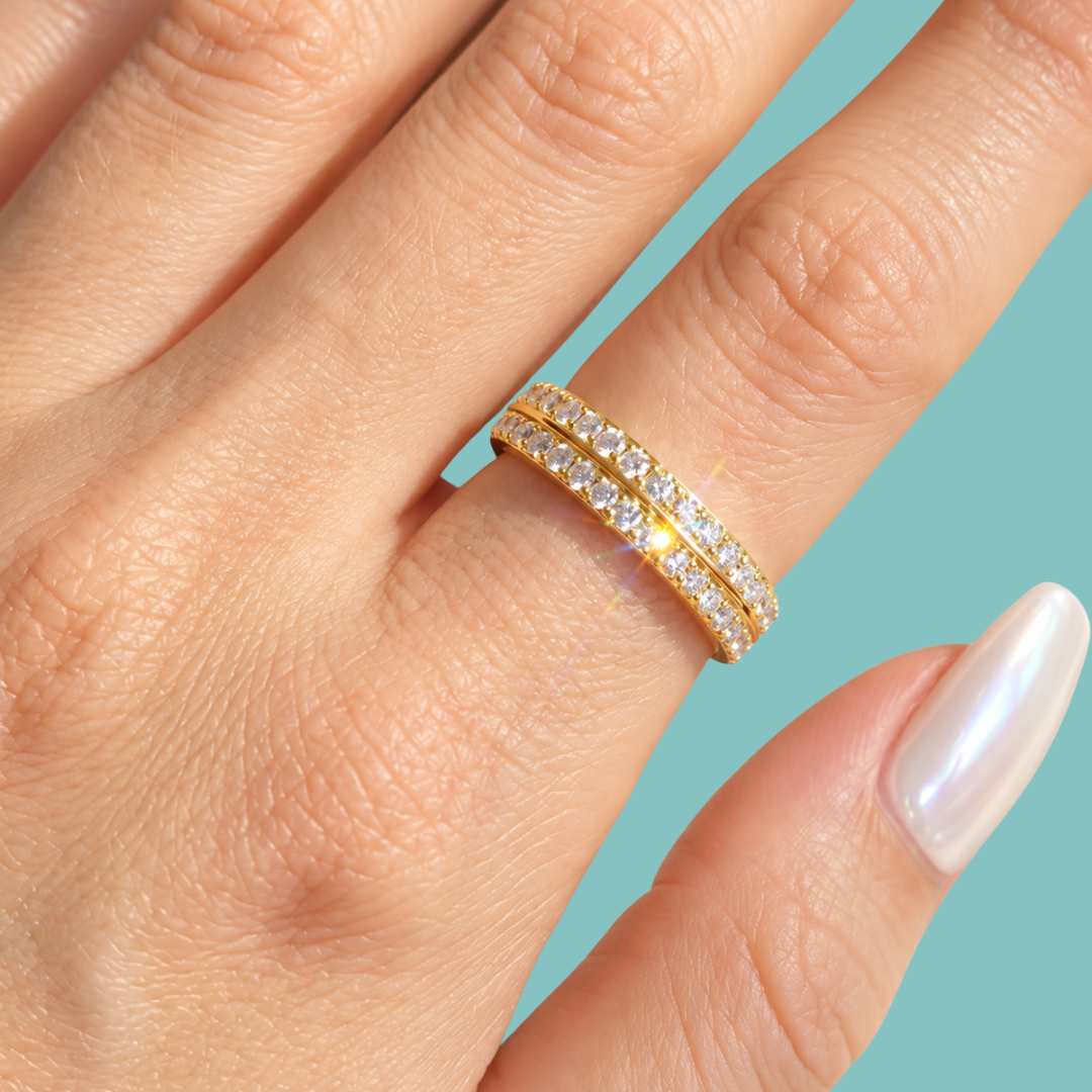 A close-up of a hand wearing a double-row yellow gold eternity ring set with clear pavé gemstones, set against a solid light blue background.