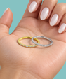 Two slim eternity rings, one yellow gold and one silver, both set with small clear stones, displayed on an open hand with manicured nails against a solid teal background.