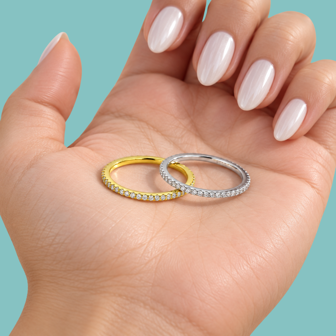 Two slim eternity rings, one yellow gold and one silver, both set with small clear stones, displayed on an open hand with manicured nails against a solid teal background.