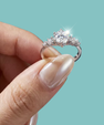 A close-up of a hand holding a silver-toned ring with a sparkling clear center stone and smaller side stones, set against a solid light aqua background.