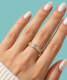 A close-up of a hand wearing a thin yellow gold eternity ring set with small clear stones, against a solid light blue background.