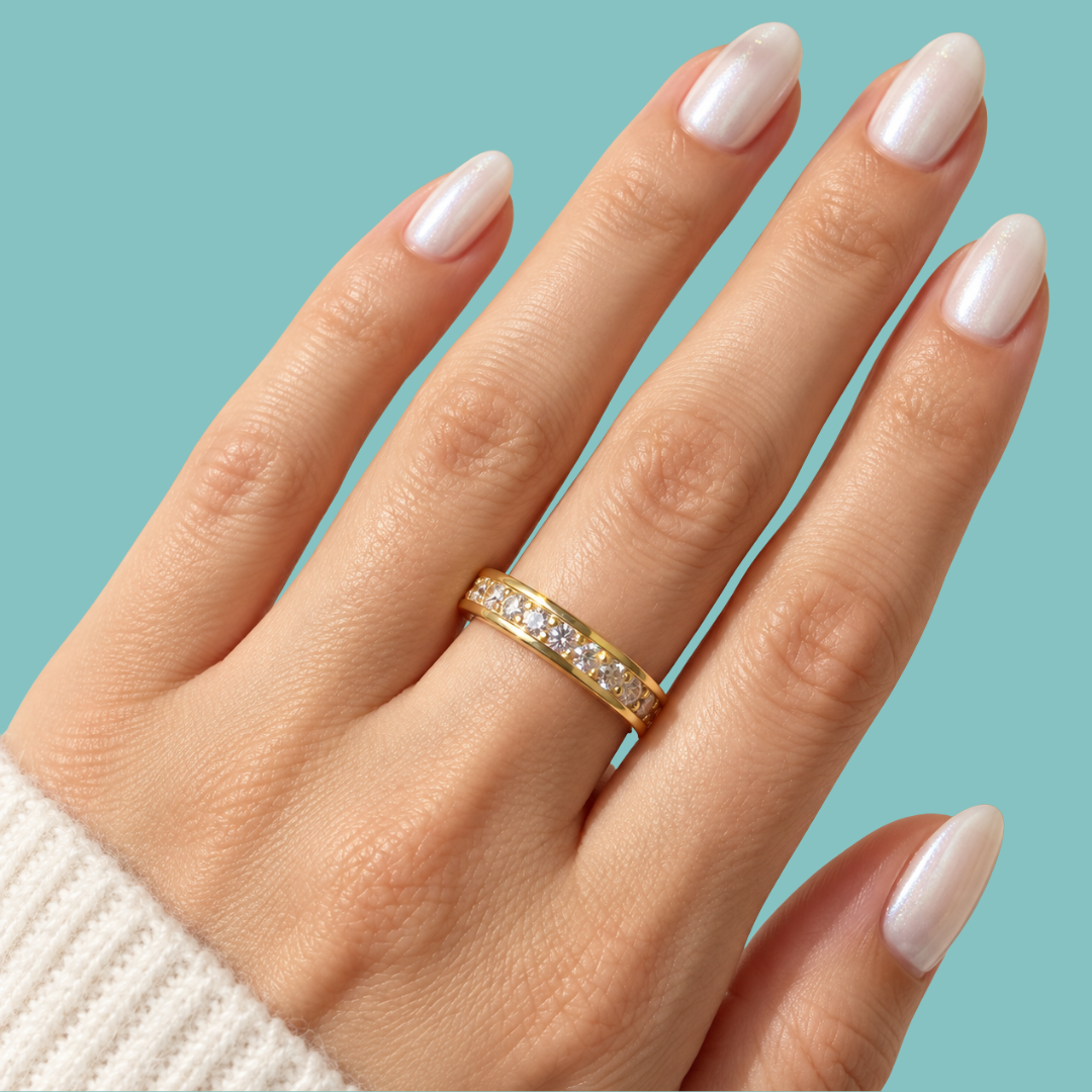 A close-up of a hand wearing a thin yellow gold eternity ring set with small clear stones, against a solid light blue background.