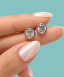 A close-up of a hand with pale pink manicured nails holding a pair of round bezel-set stud earrings with sparkling clear stones on a solid light aqua background.