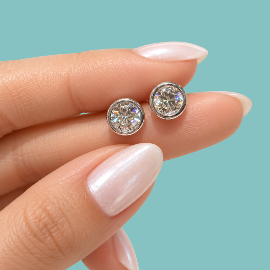 A close-up of a hand with pale pink manicured nails holding a pair of round bezel-set stud earrings with sparkling clear stones on a solid light aqua background.