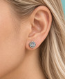 A close-up of a woman's ear wearing a round stud earring with a silver-toned halo setting and clear gemstones, against a solid teal background.