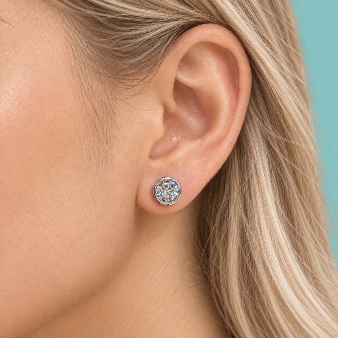 A close-up of a woman's ear wearing a round stud earring with a silver-toned halo setting and clear gemstones, against a solid teal background.