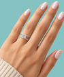 A close-up of a hand with pale pink manicured nails wearing a silver engagement ring with a round-cut center stone and a matching thin band, set against a solid light blue background.
