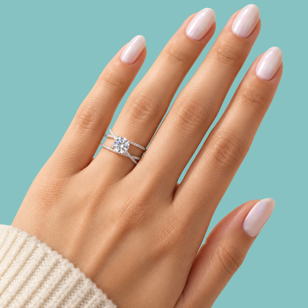 A close-up of a hand with pale pink manicured nails wearing a silver engagement ring with a round-cut center stone and a matching thin band, set against a solid light blue background.