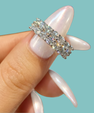A close-up of a hand with long, glossy pale pink manicured nails holding a silver band ring set with multiple clear round-cut gemstones, against a solid light aqua background.