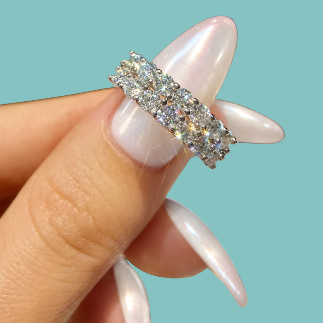 A close-up of a hand with long, glossy pale pink manicured nails holding a silver band ring set with multiple clear round-cut gemstones, against a solid light aqua background.