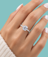 A close-up of a hand wearing a silver-toned solitaire engagement ring with a round clear center stone and small side stones, set against a solid teal background.