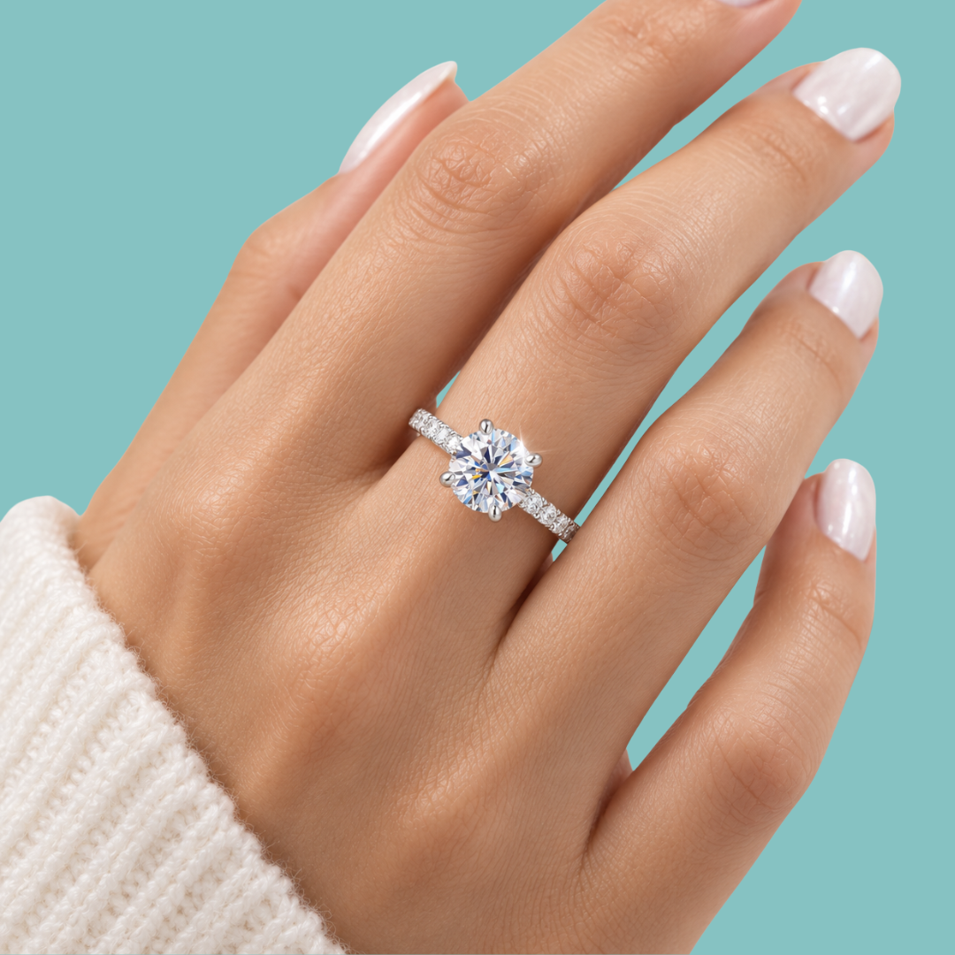 A close-up of a hand wearing a silver-toned solitaire engagement ring with a round clear center stone and small side stones, set against a solid teal background.