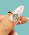 A gold solitaire ring with a round clear gemstone set in a prong setting, shown worn on a hand with long, glossy white manicured nails against a solid light blue background.