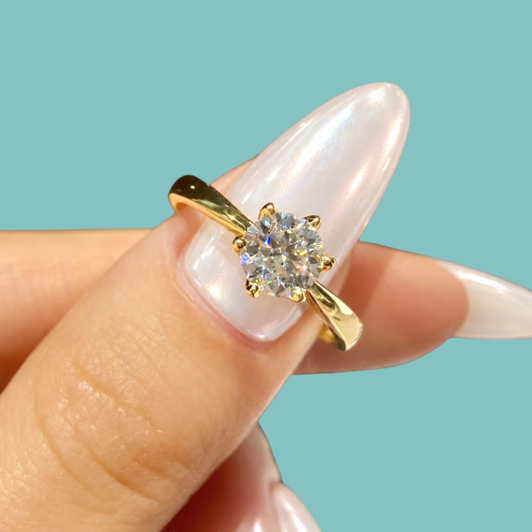 A gold solitaire ring with a round clear gemstone set in a prong setting, shown worn on a hand with long, glossy white manicured nails against a solid light blue background.