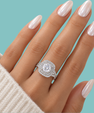 A close-up of a hand with long, glossy pale pink nails wearing a silver-toned halo ring with a large round clear center stone and smaller surrounding stones, set against a solid light aqua background.