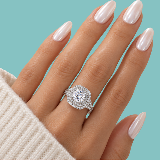 A close-up of a hand with long, glossy pale pink nails wearing a silver-toned halo ring with a large round clear center stone and smaller surrounding stones, set against a solid light aqua background.
