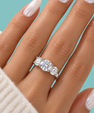 A close-up of a hand wearing a silver three-stone diamond ring, with the fingers resting against a solid teal background.
