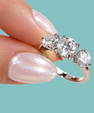 A close-up of a hand wearing a silver three-stone diamond ring, with the fingers resting against a solid teal background.