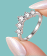 A silver-toned ring with five clear round-cut stones arranged in a curved row, held between two manicured fingernails against a solid light aqua background.