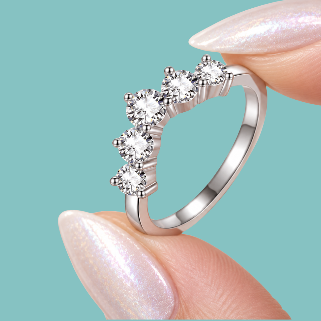 A silver-toned ring with five clear round-cut stones arranged in a curved row, held between two manicured fingernails against a solid light aqua background.