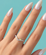 A close-up of a hand with long, glossy white manicured nails wearing a silver V-shaped ring set with small clear stones, against a solid light blue background.