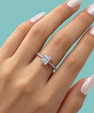 A close-up of a hand with pale pink manicured nails wearing a silver engagement ring with a large square-cut clear center stone and a pavé band, set against a solid light blue background.