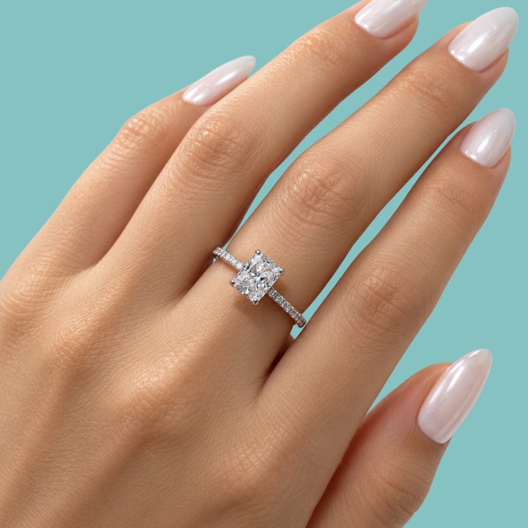 A close-up of a hand with pale pink manicured nails wearing a silver engagement ring with a large square-cut clear center stone and a pavé band, set against a solid light blue background.